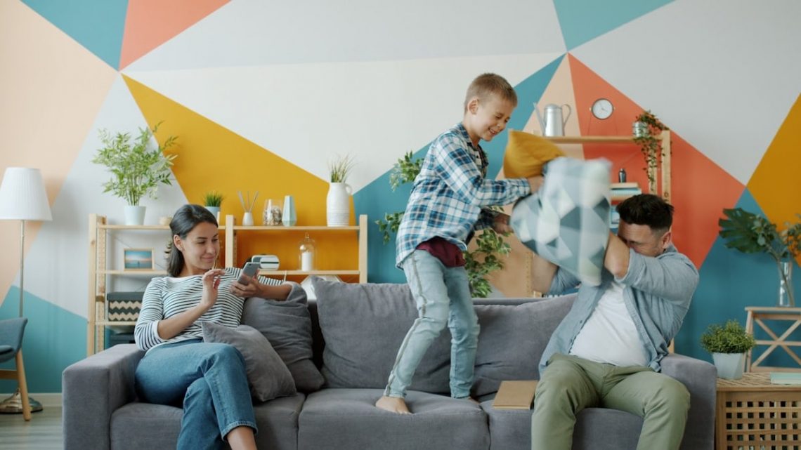 Father and son active people are enjoying pillow fight laughing while mother is using smartphone relaxing on couch at home. Family and fun concept.