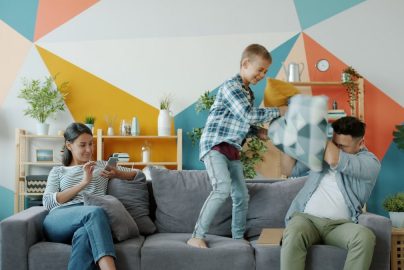 Father and son active people are enjoying pillow fight laughing while mother is using smartphone relaxing on couch at home. Family and fun concept.
