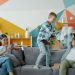 Father and son active people are enjoying pillow fight laughing while mother is using smartphone relaxing on couch at home. Family and fun concept.